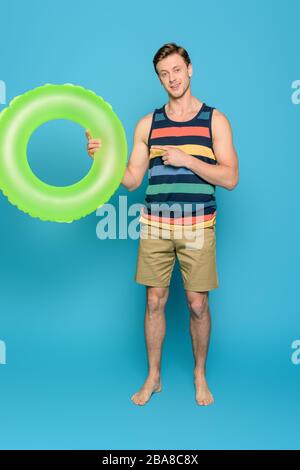 Lächelnder Mann in gestreifter Singlet und Shorts, die mit dem Finger auf den Schwimmring auf blauem Hintergrund zeigen Stockfoto
