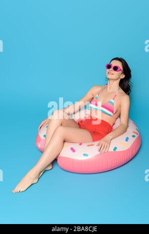 Glückliche Frau in Sonnenbrille beim Sonnenbaden auf aufblasbarem Ring auf blauem Hintergrund Stockfoto