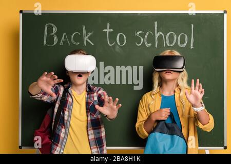 Schockierte Schulkinder mit Headsets von Virtual Reality, die in der Nähe von Schwarzen Brett mit Schriftzug "Back to School" stehen Stockfoto