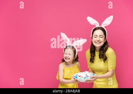 Glückliche Mutter und Tochter in Hünenähren, die Ostereier halten und isoliert auf Rosa lachen Stockfoto