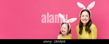 Panoramaschuss von glücklicher Mutter und Tochter in Hünenohren, die Ostereier halten und isoliert auf Rosa lachen Stockfoto