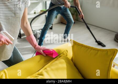 3/4-Ansicht der Frau mit Lappen und Spray in der Nähe des Sofas und eines Reinigungs-Wohnzimmers mit Staubsauger Stockfoto