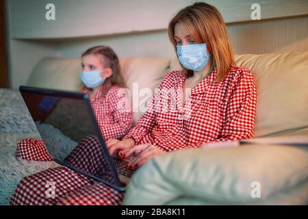 Frau in Pyjamas mit Notizbuch und Papieren, die von zu Hause aus arbeiten und Schutzmaske tragen, während ihr Kind, Tochter Computerkonsolenspiele spielen. Stockfoto