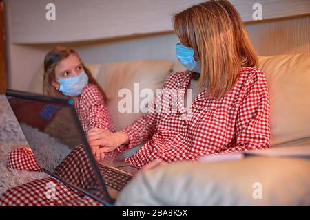 Frau in Pyjamas mit Notizbuch und Papieren, die von zu Hause aus arbeiten und Schutzmaske tragen, während ihr Kind, Tochter Computerkonsolenspiele spielen. Stockfoto