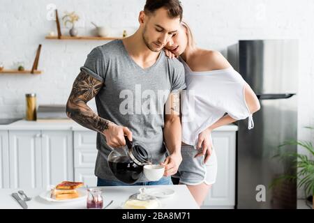 Wunderschönes Mädchen, das tätowierten Freund umarmte, der Kaffee in der Küche gießt Stockfoto