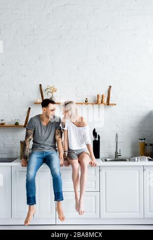 Tätowierte Paare lächeln sich an, während sie auf der Arbeitsplatte der Küche sitzen Stockfoto