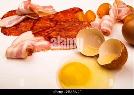 Ein zerbrochenes Hühnerei, Wurst- und Speckscheiben und Tomaten auf weißem Hintergrund. Nahaufnahme Stockfoto