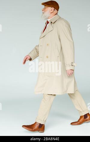 Seitenansicht des gutaussehenden Seniors im Mantel, der auf weißem Hintergrund läuft Stockfoto