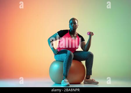 Junge afro-amerikanische und große weibliche Model-Ausbildung auf Gradientenhintergrund in Neonlicht. Trainingsübungen mit Fit Ball durchführen. Sportkonzept, gesunder Lebensstil, körperpositiv, Gleichberechtigung. Stockfoto