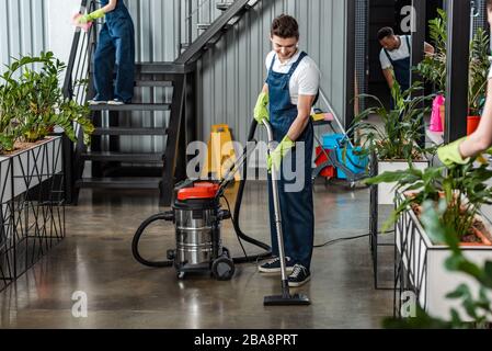Junger Reinigungsboden mit Staubsauger in der Nähe von multikulturellen Kollegen Stockfoto