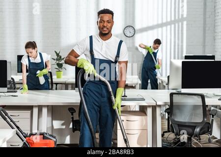 Ein gutaussehender afroamerikanischer Reiniger, der die Kamera betrachtet, während er mit Staubsauger in der Nähe von Kollegen steht Stockfoto