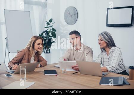 Lächelnde multikulturelle Kollegen, die in der Kreativagentur mit Gadgets sprechen und arbeiten Stockfoto