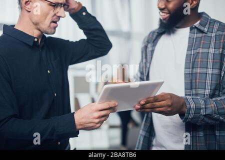 Beschneiden Sie die Ansicht, dass ein lächelnder afroamerikanischer Geschäftsmann mit seinem schockierten Kollegen über ein digitales Tablet spricht Stockfoto