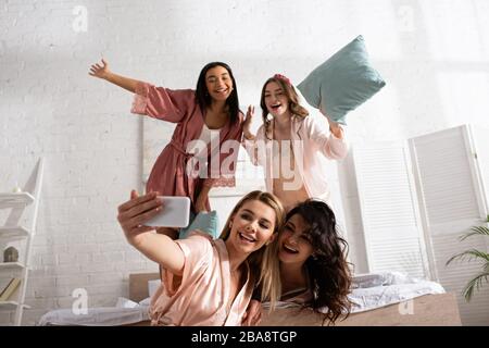 Multikulturelle Frauen lächeln, halten Kissen und nehmen selfie zusammen mit Smartphone auf dem Bett bei der Junggesellen-Party Stockfoto