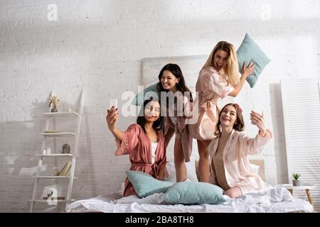 Fröhliche multikulturelle Frauen lächeln, halten Kissen und nehmen selfie mit Smartphones auf der Junggesellen-Party ins Bett Stockfoto