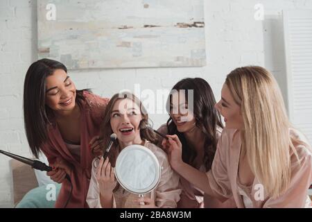 Multikulturelle Frauen, die auf der Junggesellen-Party mit Kosmetikbürsten und Spiegel und Eisenstab aufmachen Stockfoto