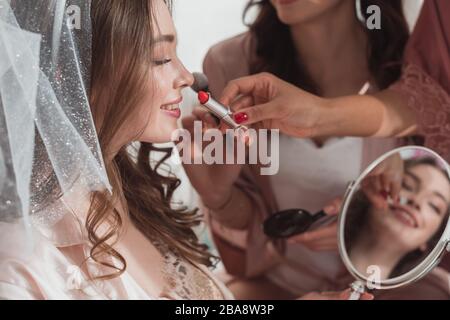 Beschnittenes Bild von multiethnischen Frauen, die Make-up mit kosmetischem Pinsel und Lippenstift auf eine Frau auf weißem Hintergrund legen Stockfoto