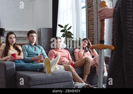 Teilweiser Blick auf den Mann in Feenking-Kostüm, der in der Nähe ernsthafter Freunde steht, die auf dem Sofa sitzen Stockfoto