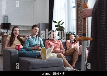 Teilweiser Blick auf den Mann in Feenking-Kostüm, der in der Nähe von lachenden Freunden steht, die auf dem Sofa sitzen Stockfoto