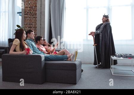 KIEW, UKRAINE - 27. JANUAR 2020: Gutaussehender Mann in Feenking-Kostüm im Gespräch mit aufmerksamen Freunden auf dem Sofa sitzen Stockfoto