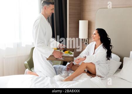 Seitenansicht des Freundes mit Frühstück im Bett für lächelnde Freundin im Hotel Stockfoto