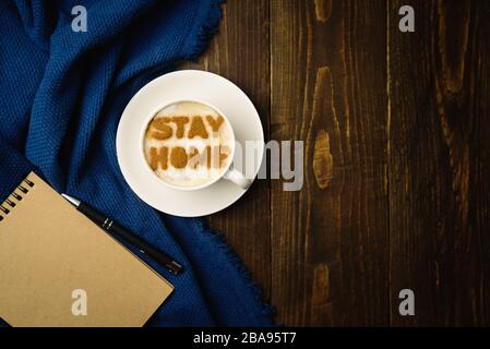 Arbeitsplatz zu Hause. Tasse Kaffee mit Text BLEIBEN SIE AUF Holztisch mit blauem Tuch ZU HAUSE. Latte Art wegen Quarantäneausbruch der Kovid-19. Stockfoto