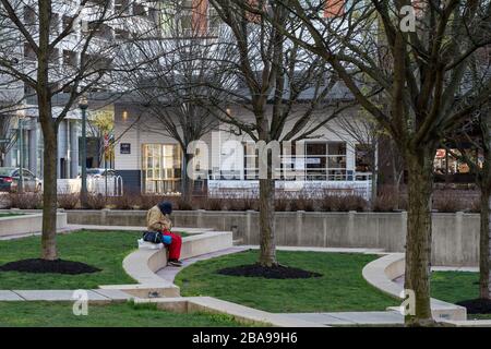 Reston, VA, USA - 26. März 2020. Ein Mann sitzt allein mit seiner Yogamatte und seiner Sporttasche im Reston Town Center Park während der Zeit von COVID - 19. Stockfoto