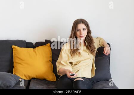 Eine Frau sitzt zu Hause auf der Couch und beobachtet Fernsehen. Sie hält eine Fernbedienung und versucht, einen interessanten Kanal zu finden. Stockfoto