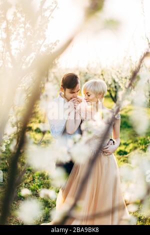 Fröhliches romantisches Paar im schönen blühenden Garten, genießen Sie den Spaziergang. Mann, der die Hand seiner hübschen Frau küsst. Blick durch die Blüte Stockfoto
