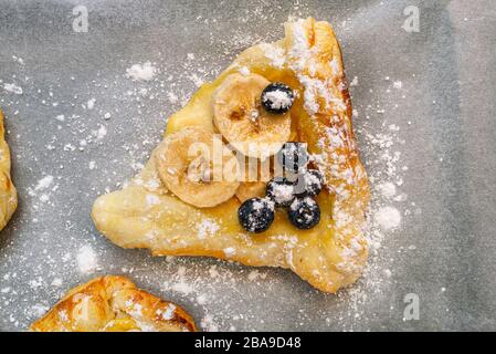 Plätzchen aus Blätterteig mit Pudding, Bananen und Blaubeeren bestreut mit Puderzucker, der auf Backpapier liegt. Stockfoto