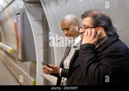 Eine Frau benutzt ihr Smartphone im Untergrund, während ein Mann telefoniert, Teil des U-Bahn-Systems London Underground Stockfoto