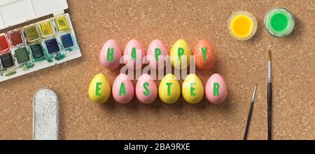 ostereier mit Botschaft HAPPY EASTER umgeben von Pinsel und Wasserfarbenboxen auf Korkhintergrund Stockfoto