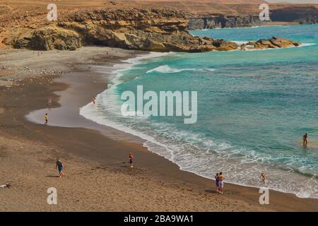 Spektakulärer schwarzer Sandstrand und Berge mit Buchten im Hintergrund in Ajuy mit Menschen, die am Ufer in Fuerteventura, Kanarieni, baden und sonnen Stockfoto