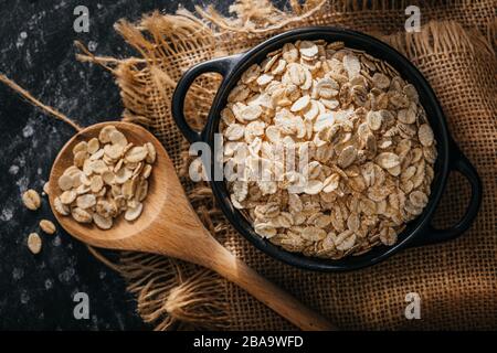 Frühstück mit Fasern für eine ausgewogene Ernährung. Haferfoto. Schwarze Schüssel und Löffel mit roher Hafer auf schwarzem Grund und rustikalem Tuch. Stockfoto