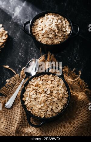 Draufsicht auf zwei schwarze Schüsseln und Löffel mit roher Haferbrei auf schwarzem Hintergrund. Frühstück mit Fasern für eine ausgewogene Ernährung. Haferfoto Stockfoto