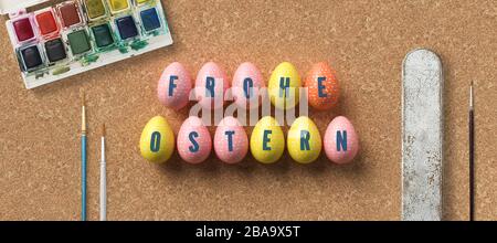 ostereier mit Botschaft HAPPY EASTER auf Deutsch umgeben von Pinsel und Wasserfarbenboxen auf Korkhintergrund Stockfoto