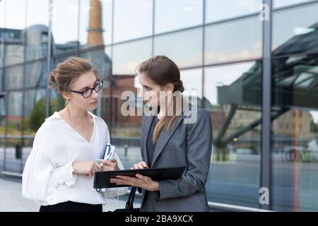 Zwei junge und schöne Geschäftsfrauen stehen vor dem Hintergrund eines Bürogebäudes. Sie diskutieren die Grafiken, die sie auf dem Tisch gesehen haben Stockfoto
