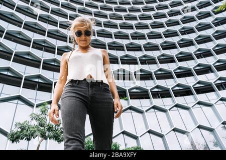 Blondes Mädchen mit Sonnenbrille und einem Geflecht, das vor einem modernen Gebäude steht, das auf die Kamera in Singapur blickt Stockfoto