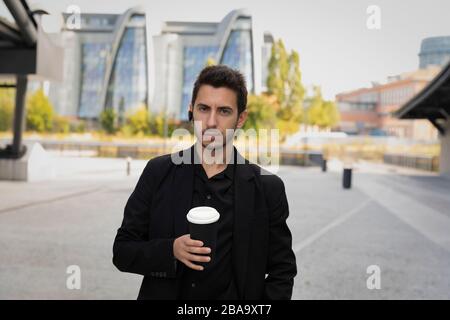 Ein junger gutaussehender Mann steht in einem schwarzen Business-Anzug auf dem Hintergrund eines Bürogebäudes. Er trinkt seinen Kaffee aus einer nachfüllbaren Tasse und nimmt Stockfoto