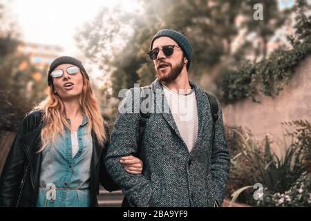 Ein paar junge Leute wachen im Garten des Einkaufszentrums draußen. Sonnenbrille und warmer Hut tragen. Stockfoto