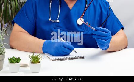 Ein männlicher Arzt in blauer Uniform sitzt an einem weißen Tisch und schreibt in einem Papier-Notizbuch, trägt sterile Handschuhe an den Händen, Konzept der Patien Stockfoto