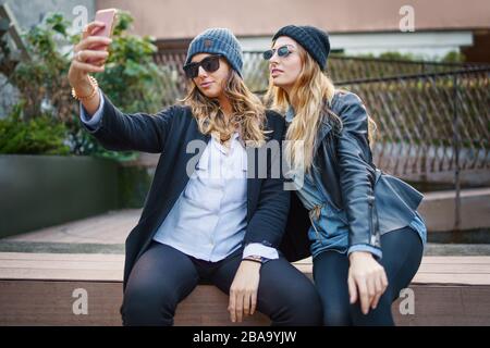 Zwei junge glückliche Mädchen, die auf einer Bank sitzen und Selfies machen. Beste Freunde, die Spaß haben, mit dem Smartphone Selbstbilder zu machen, das Hüte und Sonnenbrille trägt. Stockfoto