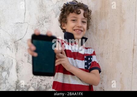 Schuljunge, die Smartphone-Bildschirm draußen vor einem texturierten Hintergrund aus Betonwand zeigen Stockfoto