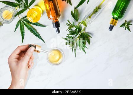 Pipettieren Sie kosmetisches CBD-Öl in weiblichen Händen auf Tischhintergrund mit Kosmetik, Creme mit Cannabis und Hanfblättern Stockfoto