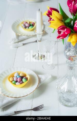 Serviert wird Ostertisch mit bunten süßen Eiern in den Fällen auf den Tellern, Kristallglas und frischen Tulpenblumen auf dem weißen Holztisch. Familienhol Stockfoto