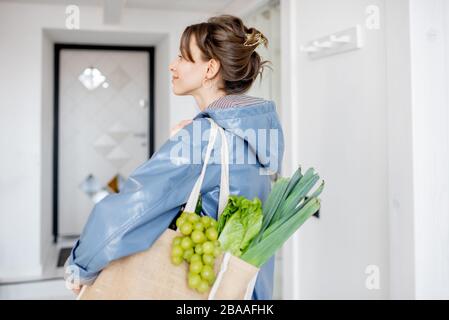 Junge Frau in blauem Mantel kommt mit Einkaufstasche voller frischem Gemüse und Grüns nach Hause und läuft auf dem Flur Stockfoto