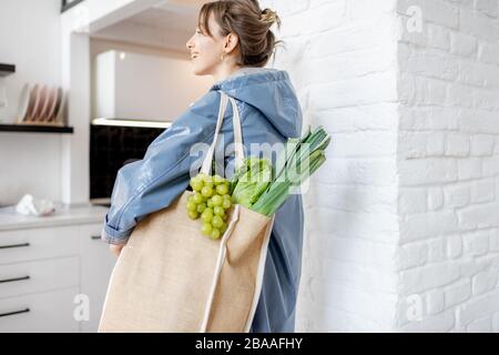 Junge Frau in blauem Mantel kommt mit Einkaufstasche voller frischem Gemüse und Grüns nach Hause und läuft auf der Küche Stockfoto