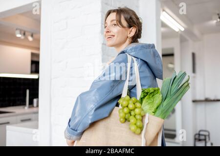 Junge Frau in blauem Mantel kommt mit Einkaufstasche voller frischem Gemüse und Grüns nach Hause und läuft auf der Küche Stockfoto