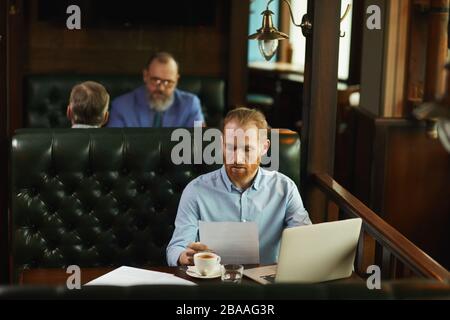 Viel reifer Mann sitzt am Tisch und arbeitet mit Dokumenten und einem Laptop, während er Kaffee im Café trinkt Stockfoto