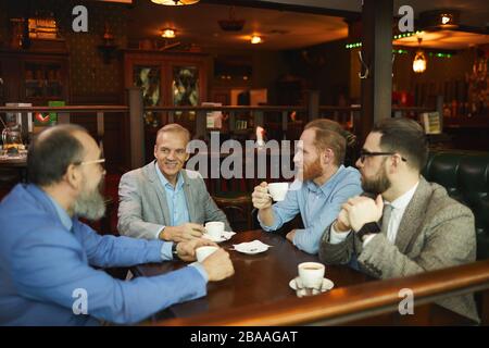 Gruppe von Geschäftsleuten in Anzügen, die mit Tassen Kaffee am Tisch sitzen und sich bei einem Treffen im Café miteinander unterhalten Stockfoto